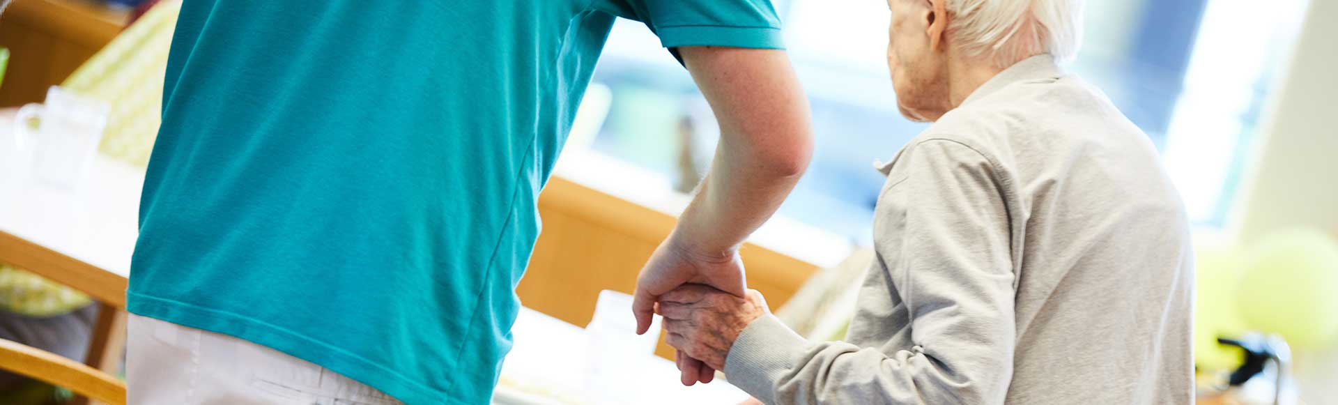 Caregiver offering support to an elderly person in a facility with commercial laundry equipment like commercial tumble dryers and automatic ironing machines.