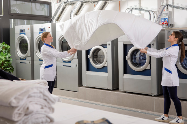 Two women in uniforms shaking out a large white duvet in a commercial laundry facility, with industrial washing machines in the background.