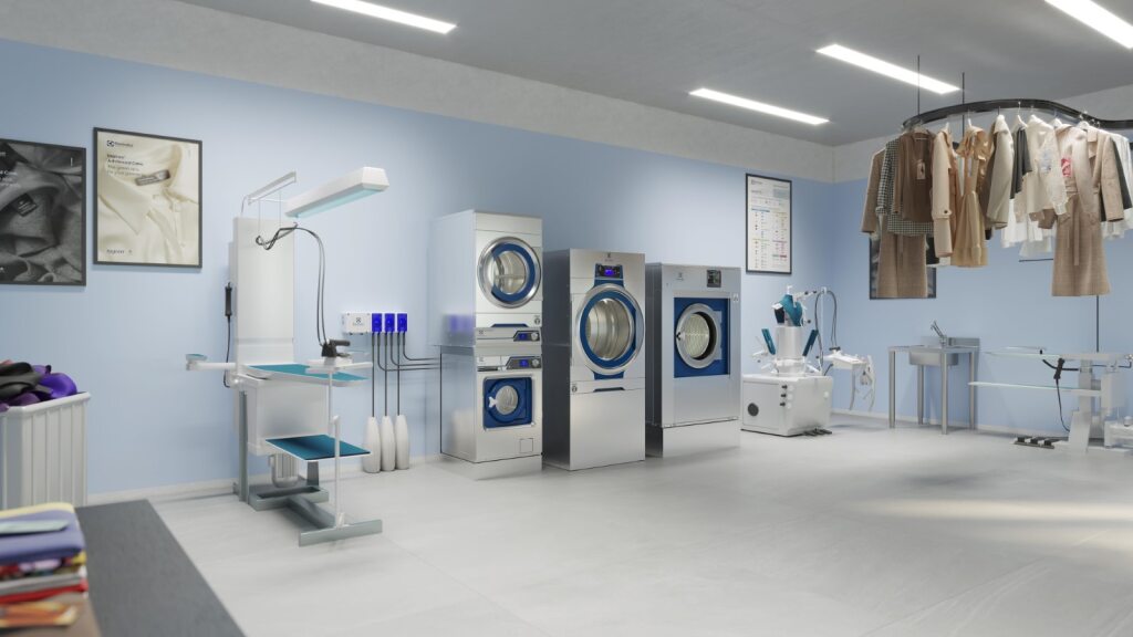 A professional laundry or dry cleaning room is shown, with several industrial-sized washing machines and dryers lined up against a light blue wall. The machines are white and stainless steel, with blue accents around the doors. On the left, there is a specialized ironing or pressing station with a large lamp. In the foreground on the left, there is a pile of folded laundry. On the right, a circular conveyor rack holds a variety of garments, including shirts and jackets, suggesting a dry-cleaning service. The room is brightly lit with long fluorescent lights on the ceiling and appears clean and modern.