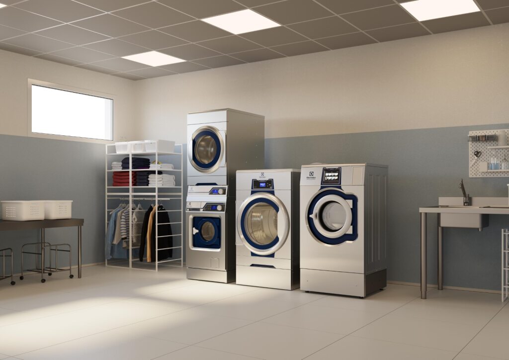 Professional laundry room with three stainless steel Electrolux Professional machines: a stacked dryer and washer unit, a front-loading washer, and a front-loading dryer; on the left, shelves with hanging clothes and folded laundry; on the right, a stainless steel sink and wall-mounted storage panel.
