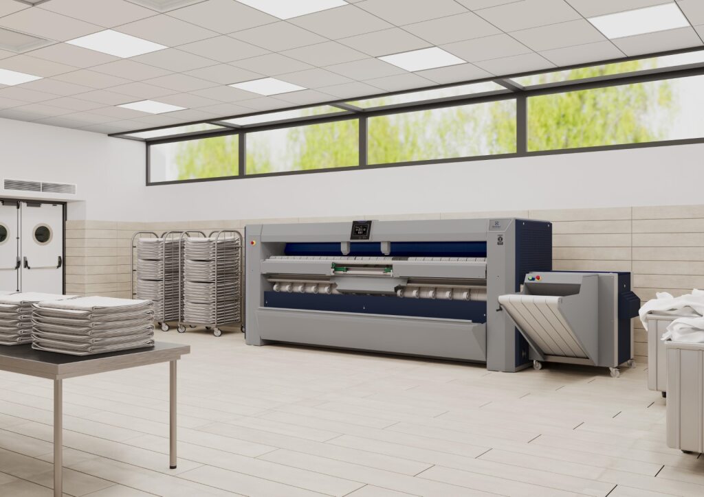Modern industrial laundry facility with a large flatwork ironer machine, neatly stacked folded linens on a table, and several metal laundry carts in a clean, well-lit room with large windows.