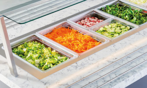 Salad bar with a variety of fresh ingredients including shredded carrots, sliced radishes, cucumbers, and leafy greens, displayed on a marble countertop.
