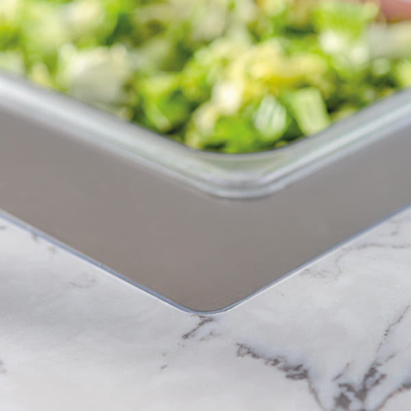 Close-up of a marble countertop with a stainless steel tray filled with chopped green vegetables in soft focus, emphasizing the smooth surface and modern design.