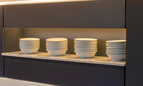 Stacks of white ceramic bowls neatly arranged on a modern marble shelf with under-cabinet lighting, showcasing minimalist kitchen shelving design.