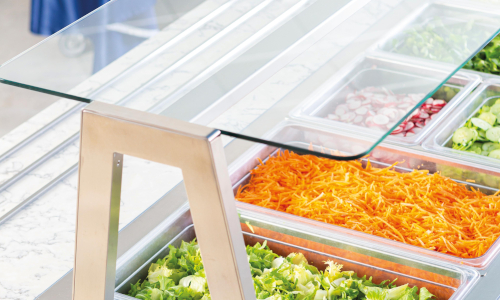 Salad bar with glass cover showing trays of shredded carrots, chopped lettuce, and other salad ingredients on a marble countertop.