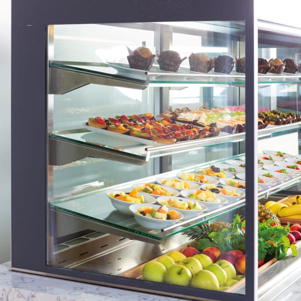 A glass display case featuring assorted desserts and fresh fruits, including muffins, layered pastries, fruit salads, apples, and bananas.