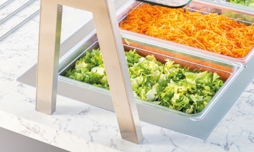 Salad bar with glass cover showing trays of shredded carrots, chopped lettuce, and other salad ingredients on a marble countertop.